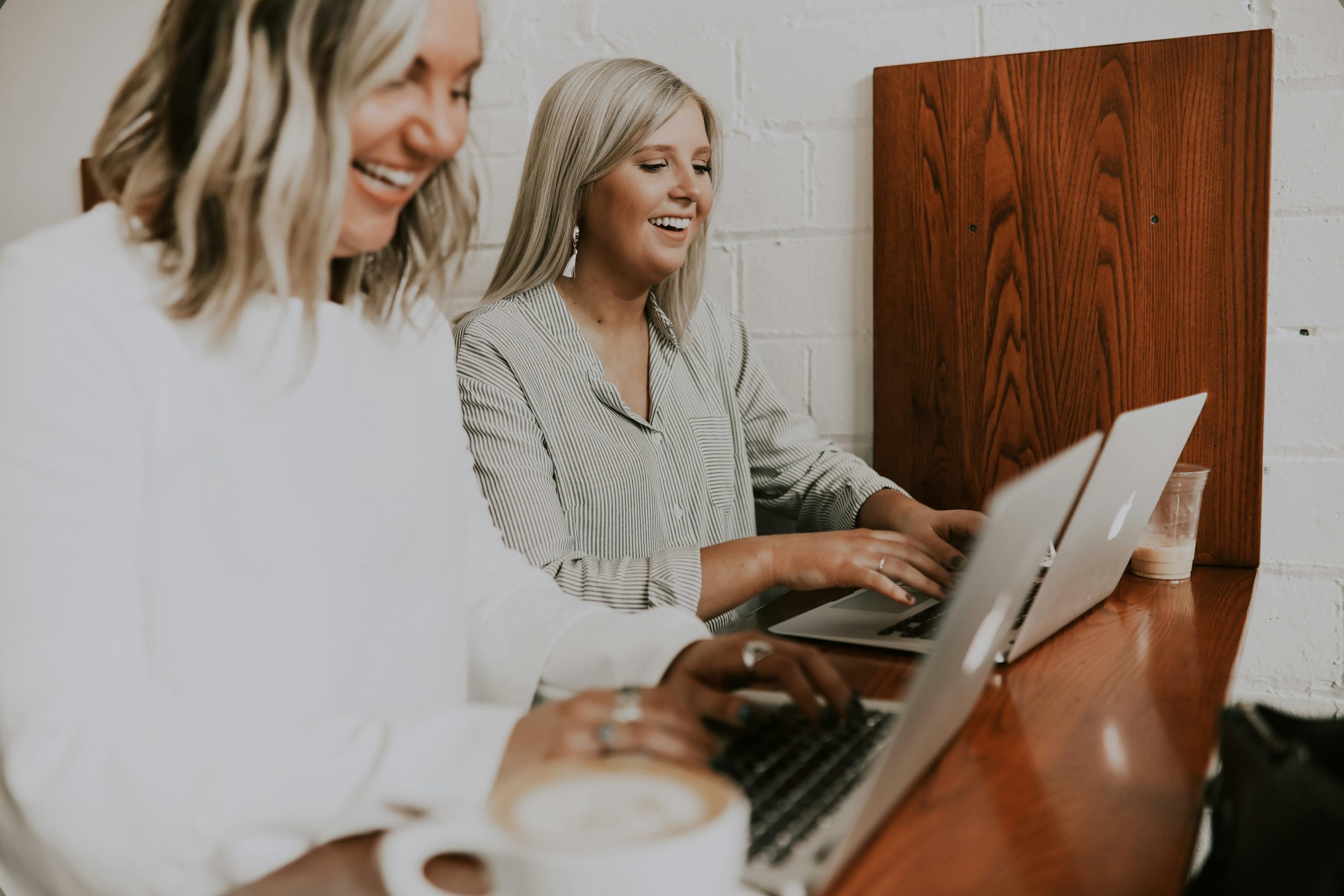 Deux femmes souriantes assises côte à côte dans un café, travaillant sur leurs ordinateurs portables, avec une tasse de café au premier plan.