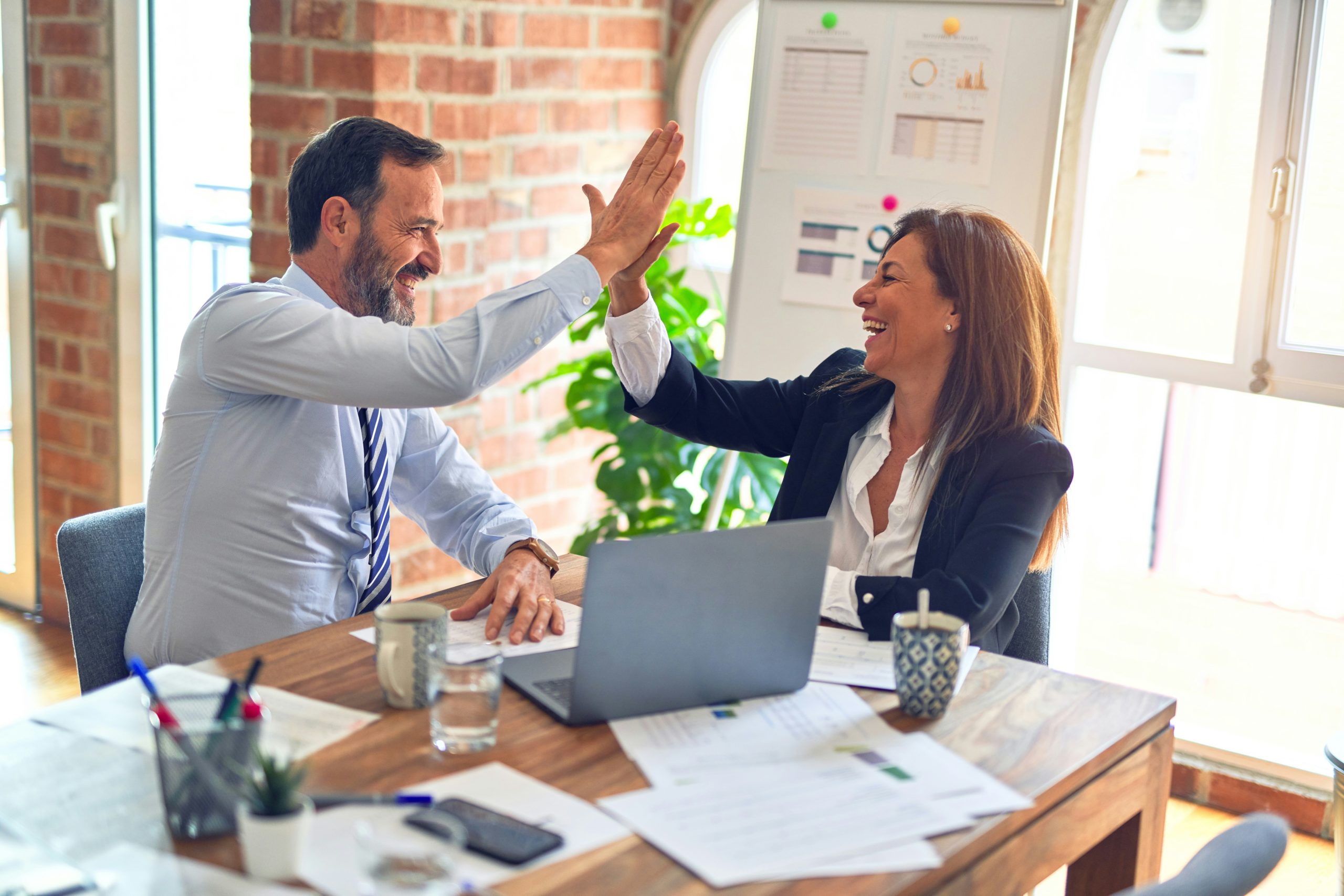 Un homme et une femme d’affaires souriants échangent un high-five lors d’une réunion autour d’une table avec un ordinateur portable, des documents et des tasses.