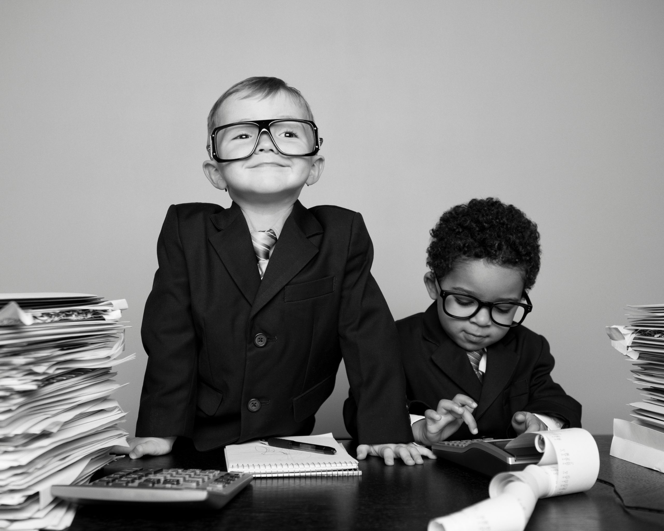 Deux enfants en costume d’affaires portant des lunettes, assis à un bureau avec piles de papiers, une calculatrice et un carnet. L’un sourit en regardant devant, l’autre utilise une calculatrice. @RichVintage