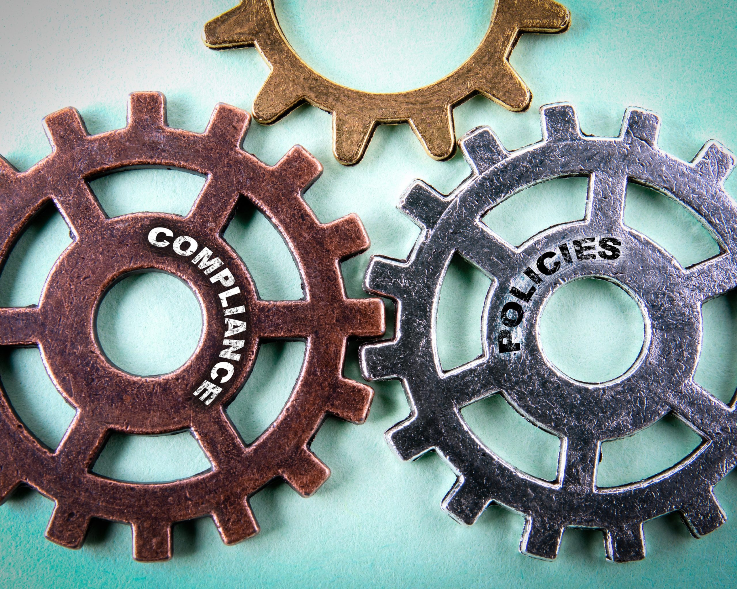 Trois engrenages métalliques imbriqués avec les mots "COMPLIANCE" et "POLICIES" inscrits sur deux d’entre eux, symbolisant la conformité et les politiques d’entreprise. @tumsasedgars
