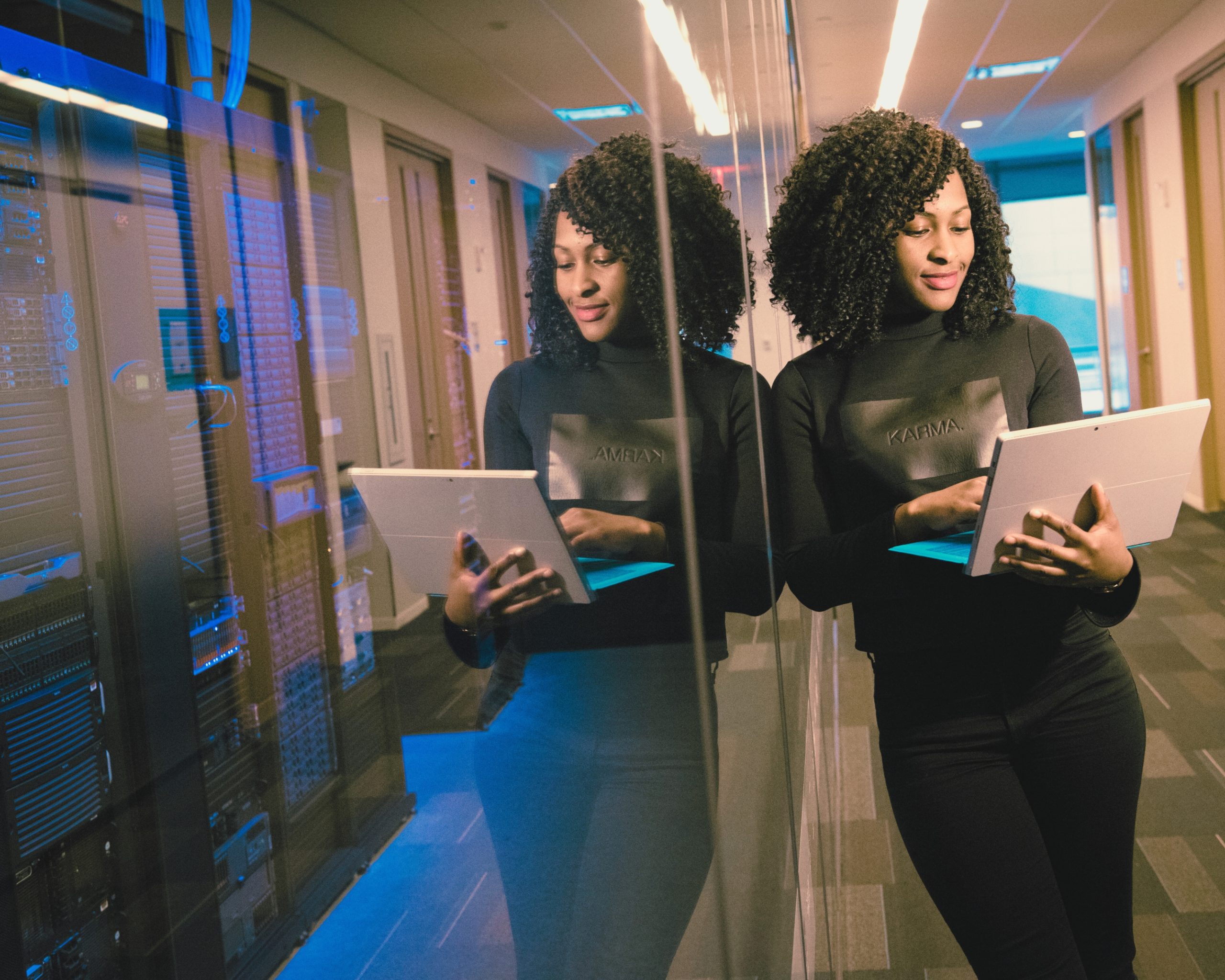 Femme noire tenant une tablette numérique, debout dans un couloir de centre de données avec serveurs, reflet dans une vitre. @ChristinaMorillo