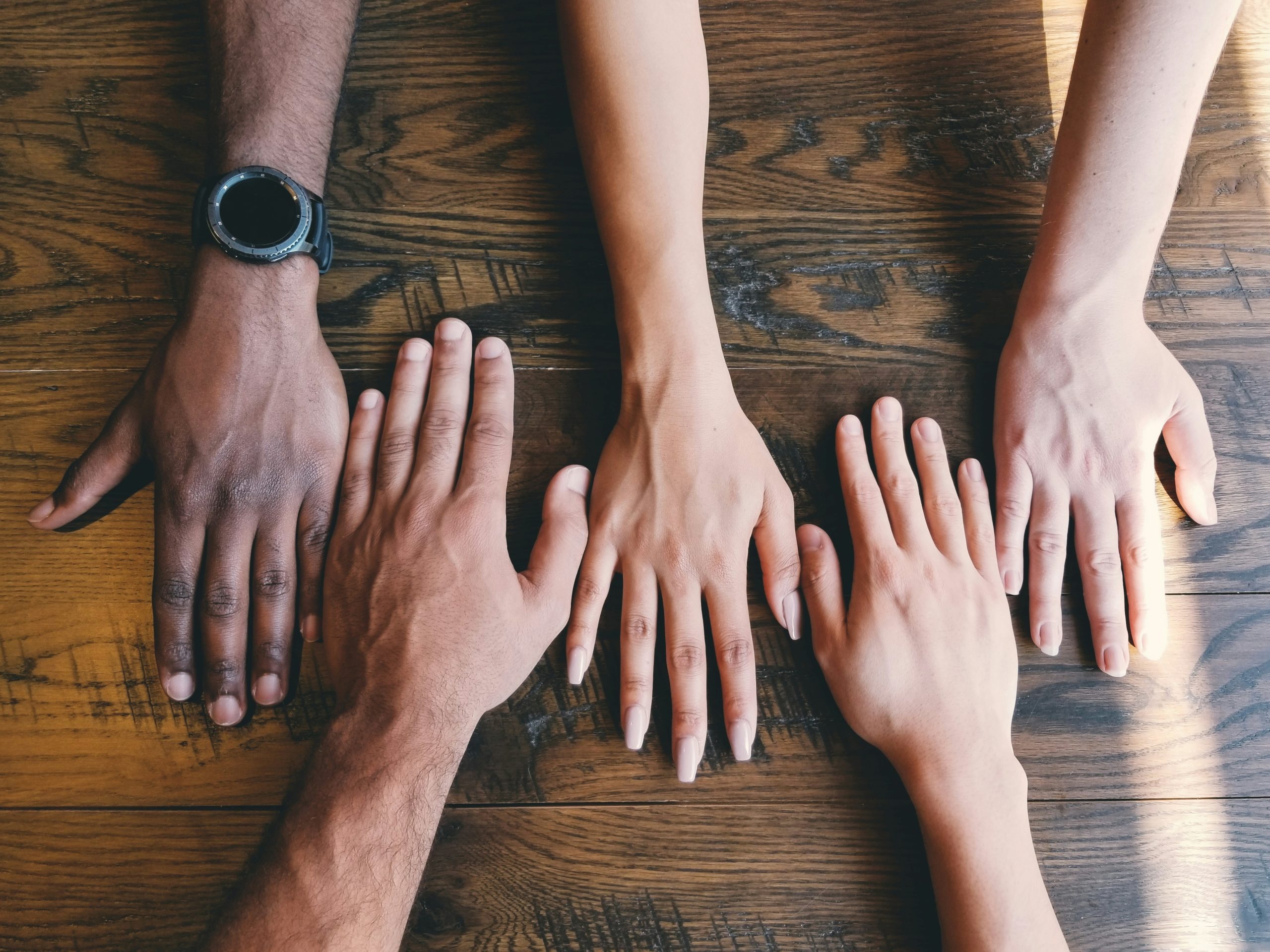 Quatre mains de personnes aux origines diverses posées à plat sur une table en bois, symbolisant l’unité et l’inclusion.