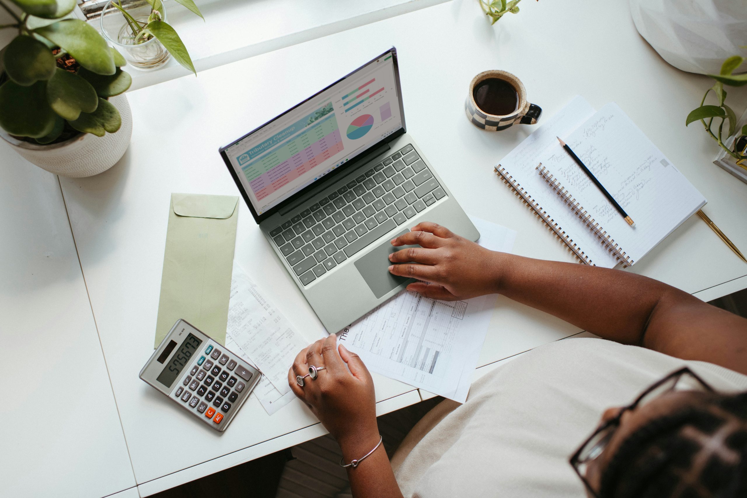 Personne travaillant sur un ordinateur portable avec des graphiques financiers à l’écran, accompagnée d’une calculatrice, de documents, d’un carnet et d’un café.